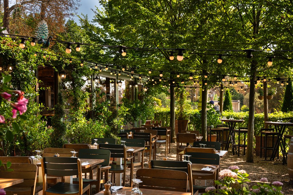 Terrasse de L’Orangerie sous les arbres, tables en bois et guirlandes lumineuses